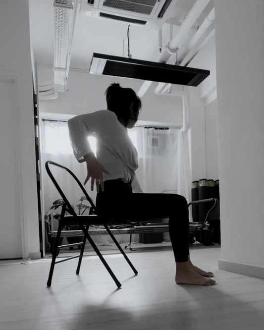 A woman in exercise attire performing a GYROKINESIS sideway arch movement on a chair, stretching her torso to the side with controlled form